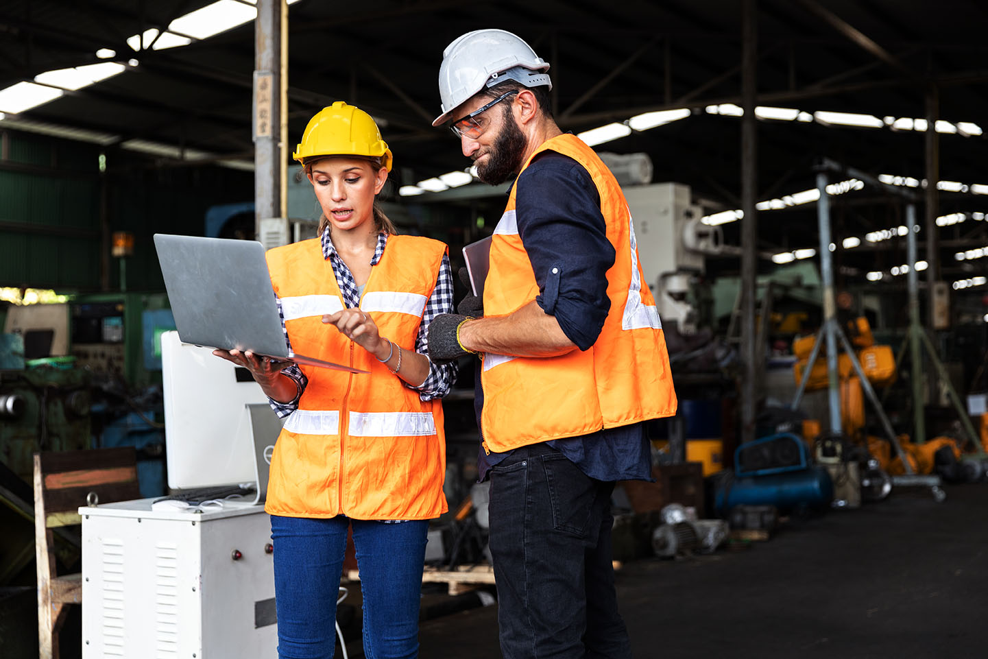 Male and female mechanical engineers or factory workers with safety gears and hard hats discuss about production project using computer laptop to plan in a warehouse