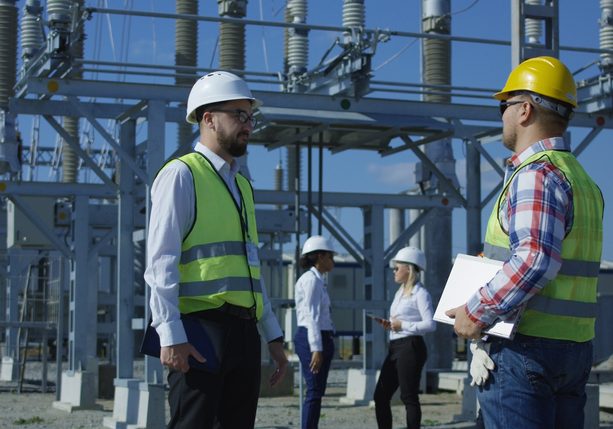 Medium shot of a group of electrical workers in safety vests talking to each other against the background of a transformer station
